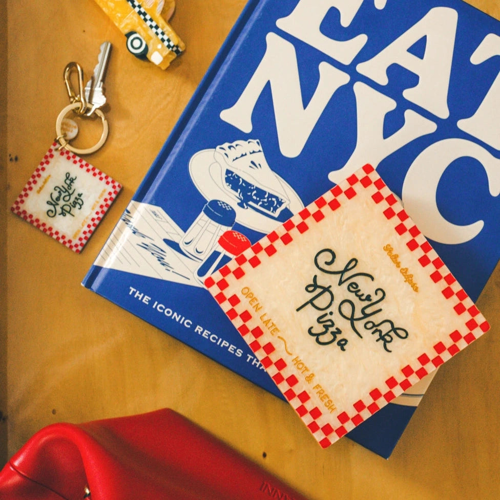 a collection of objects on a desk, including a keychain, a taxi-shaped hair clip, a cookbook reading 'eat nyc' and a red and white pizza box-styled trinket box, all over a yellow wood background