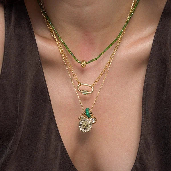 A close-up of a person wearing layered necklaces, including a gold chain with a silver and green pendant, a gold chain with a green bead accent, and a green beaded chain.