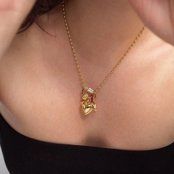 A close-up of a model wearing a delicate gold chain necklace with a small heart-shaped pendant featuring a tiny angel and red gemstone details.