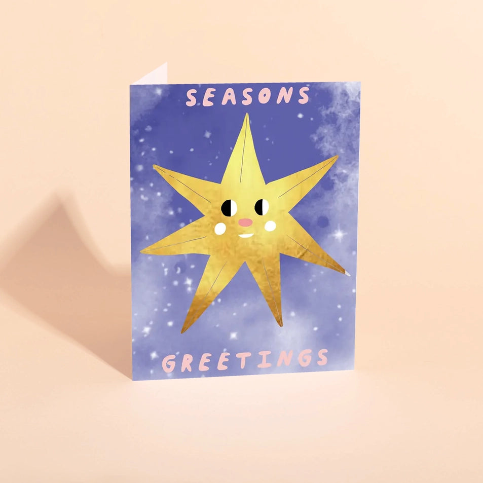 a greeting card with a blue background with white snow-like accents and a large illustration of a gold star with a face and text reading seasons greetings