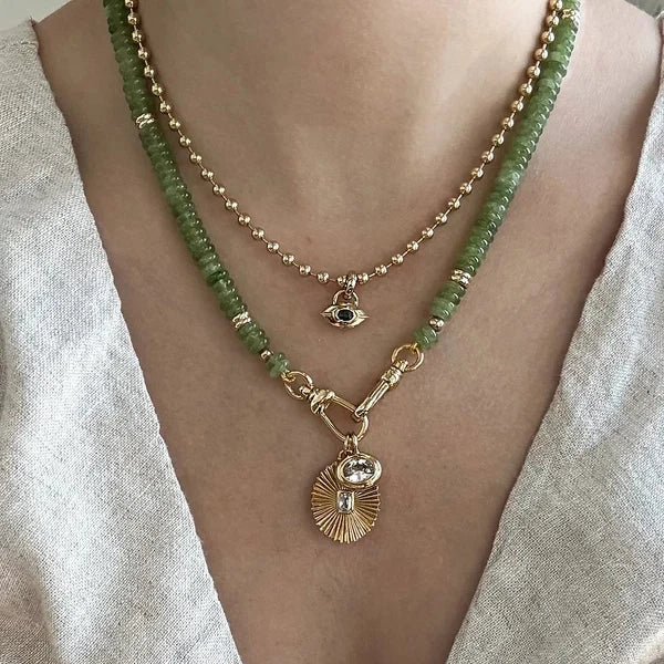 A close-up of a person wearing a layered necklace with a green beaded strand, a gold beaded chain, a gold eye charm, and a gold sunburst pendant with a clear stone.