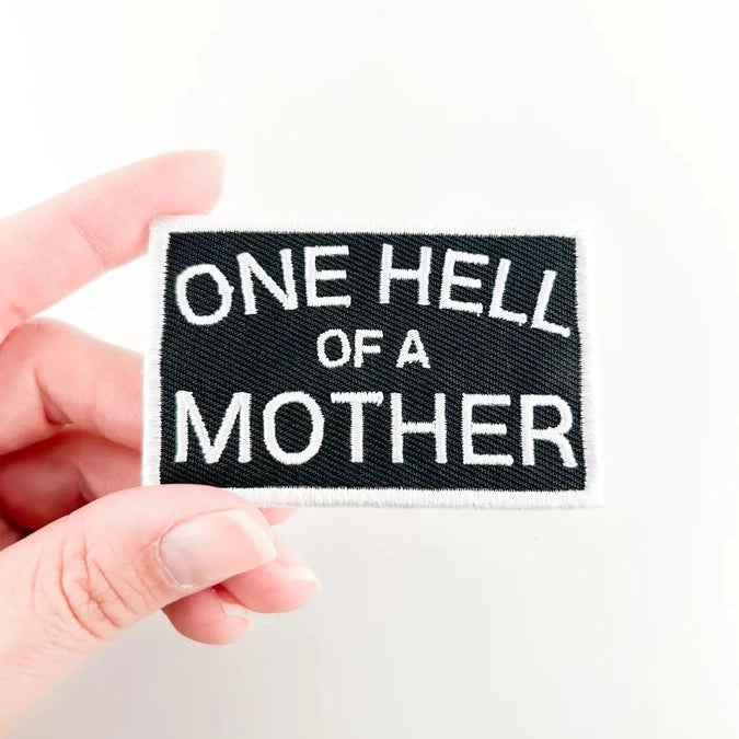 a black and white iron on patch with text reading one hell of a mother held by a hand over a white background