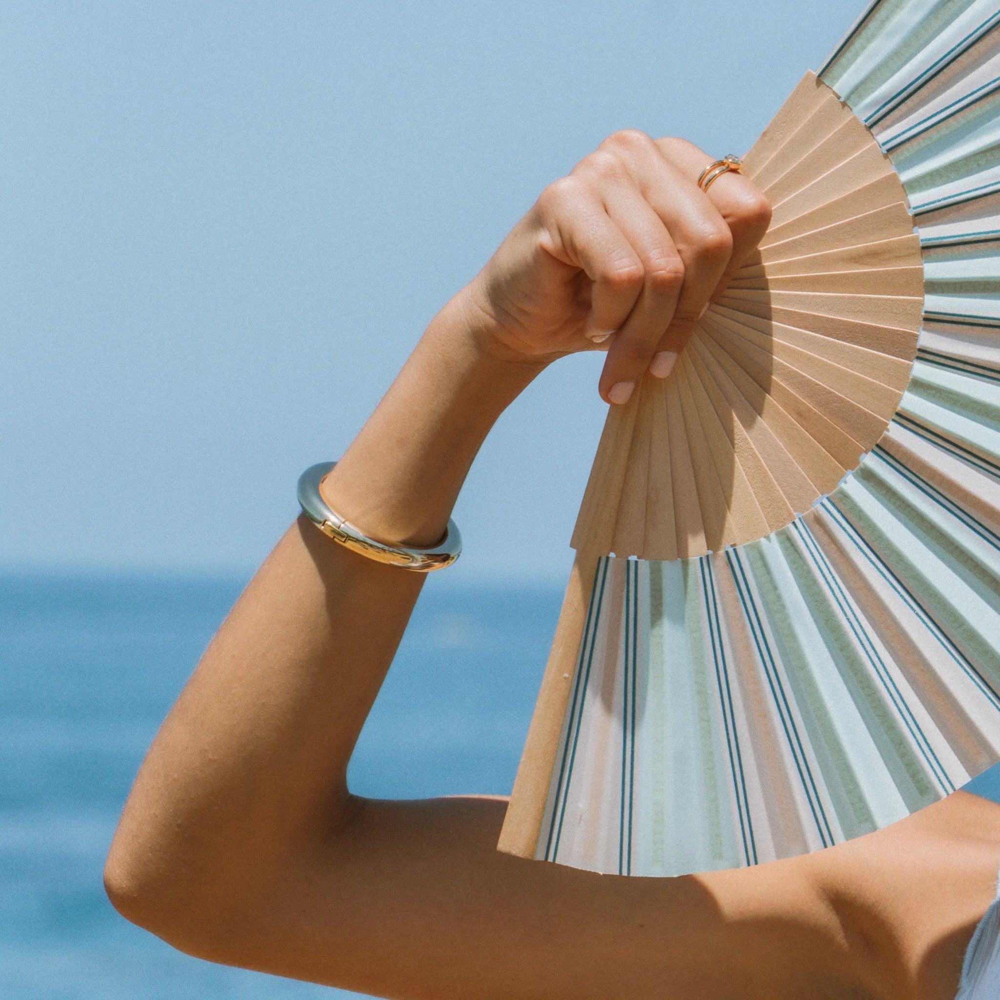 a woman wearing a thick gold bangle and holding a paper fan with an ocean background