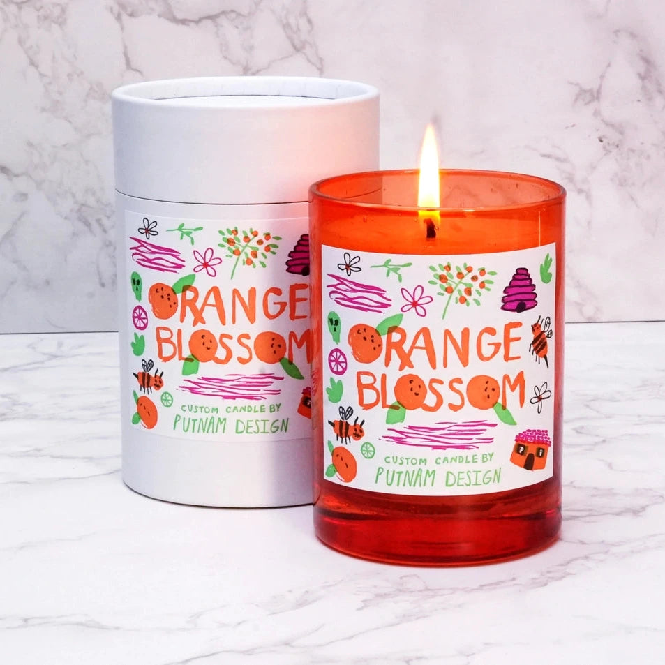 an orange candle with a white label with orange text reading orange blossom surrounded by handdrawn illustrations all over a marble background