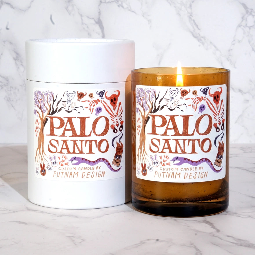 a brown lit candle with a white label with text reading palo santo surrounded by halloween motif illlustrations over a marble background