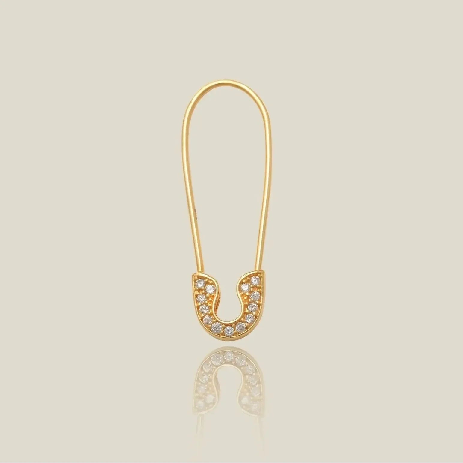 A gold safety-pin style earring with a horseshoe-shaped setting paved with small clear stones, shown on a light beige background with a soft reflection below.