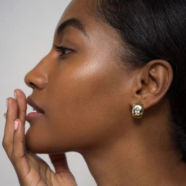 A close-up side profile of a woman wearing a small gold stud earring with a clear gemstone, set against a plain light gray background.