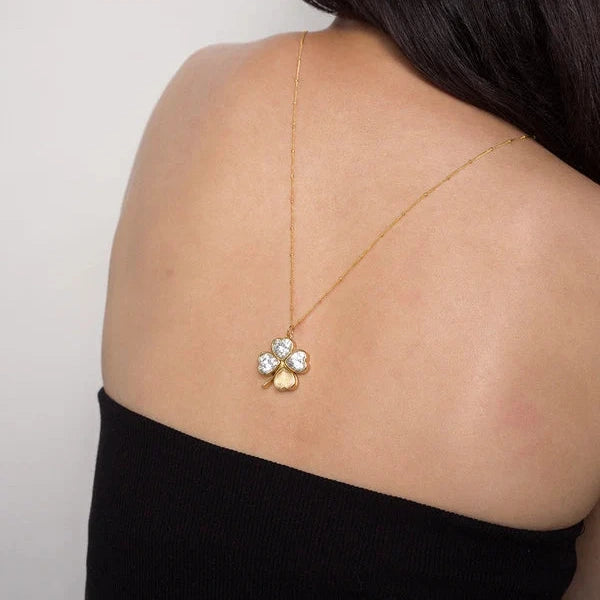 A gold necklace with a three-petal flower pendant featuring clear crystals and a small gold heart, shown on a model's back against a light gray background.
