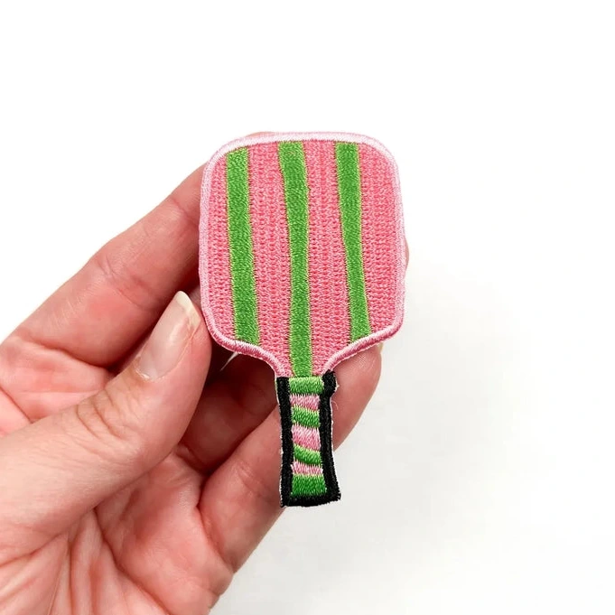 a pink and green iron on patch of a pickleball paddle held by a hand over a white background