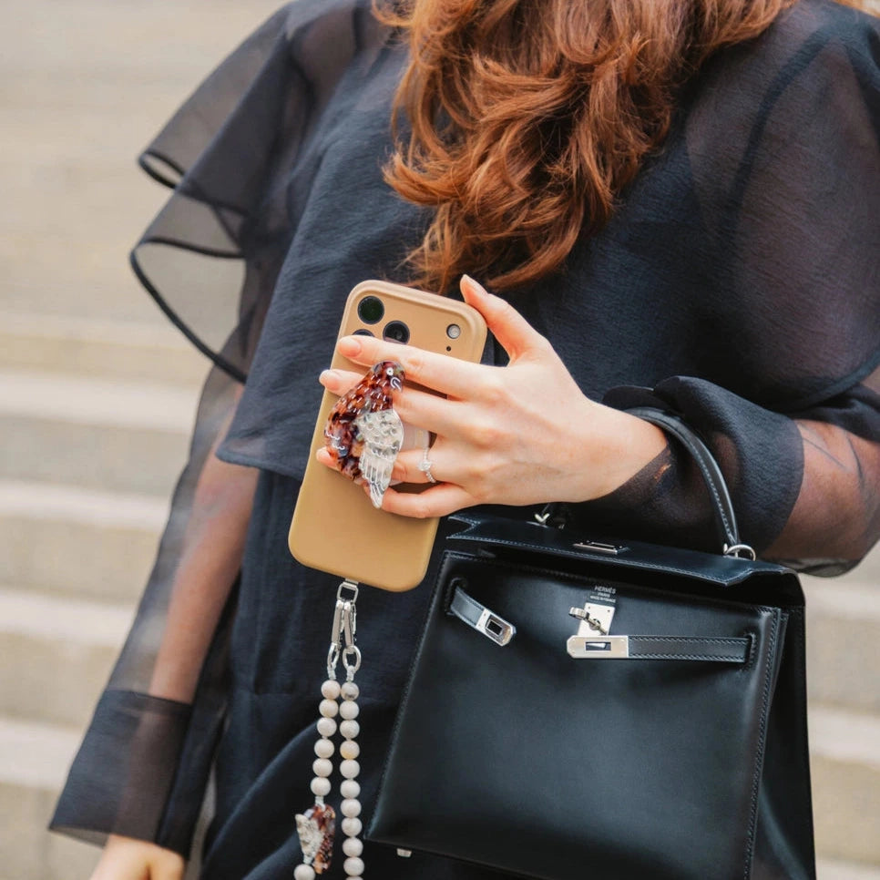 A woman in a sheer black dress carries a structured black leather handbag with silver hardware while holding a gold phone case with a pearl strap charm.
