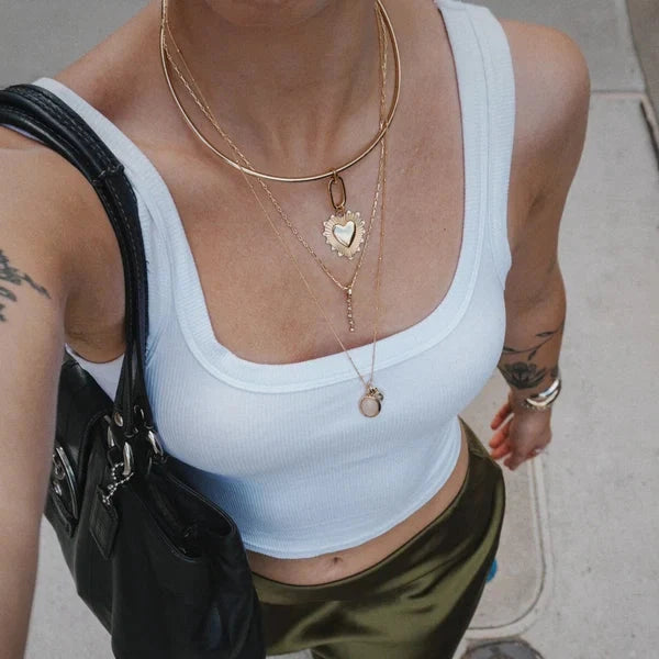 A person wearing a white ribbed scoop-neck crop top and olive green satin skirt, accessorized with layered gold necklaces, a black shoulder bag, and flip-flops on a concrete sidewalk.