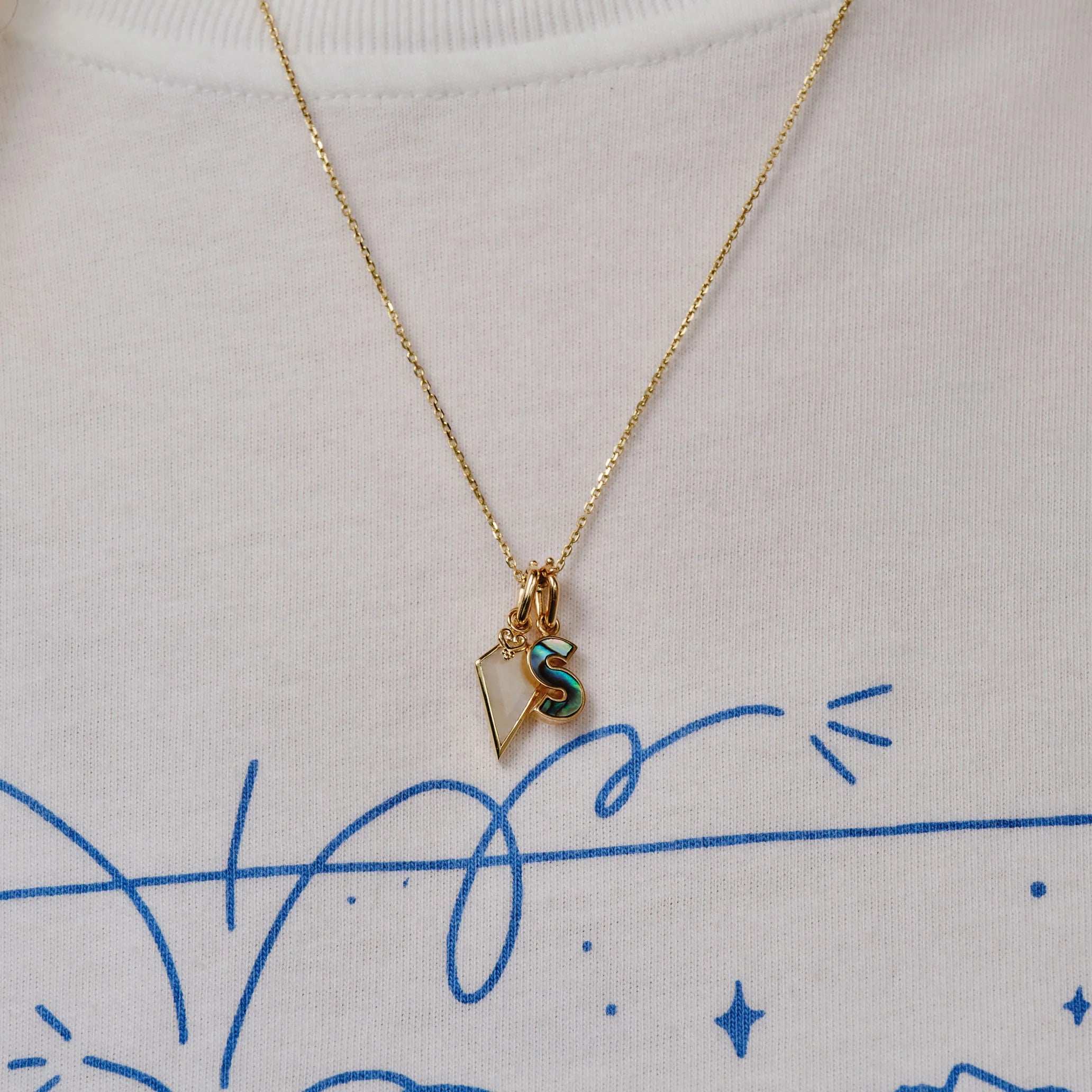 A gold chain necklace with a small initial 'S' pendant featuring a green and gold pattern, displayed on a white t-shirt with blue graphic lines.