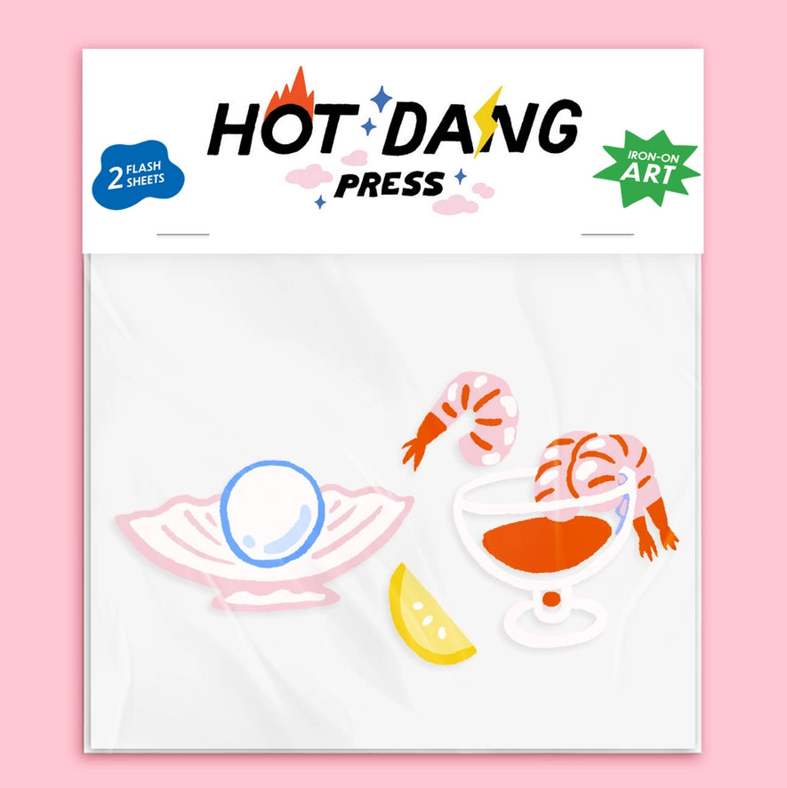 A packaged sheet of decorative stickers or decals from Hot Dang Press, featuring illustrated shrimp, a shell with a blue oval, a glass of orange drink, and a lemon slice on a white sheet against a pink background.