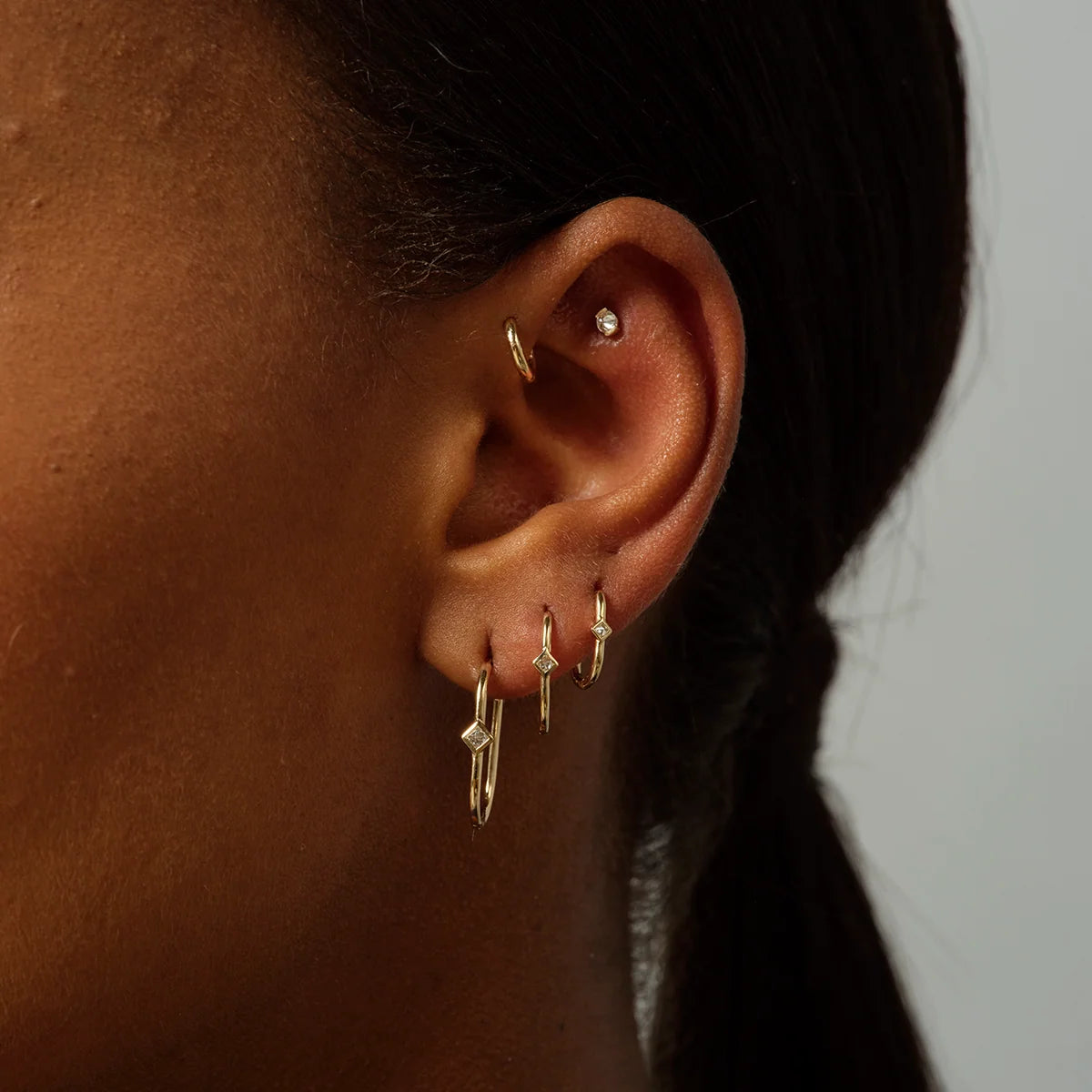 a womans ear with several earrings including three sizes of an oval shaped gold hoop with bezel set square white diamonds