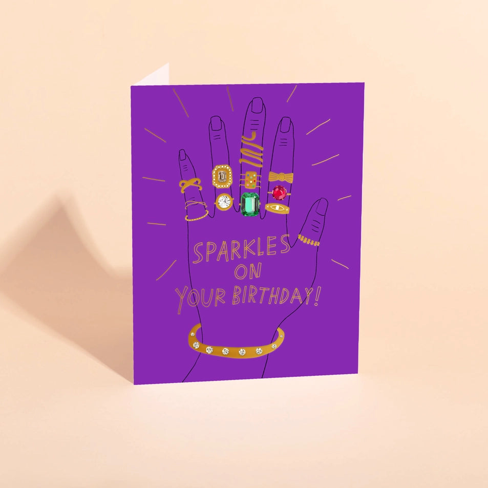 a greeting card with a purple background and an illustration of a hand wearing many gold rings overlaid with text reading sparkles on your birthday