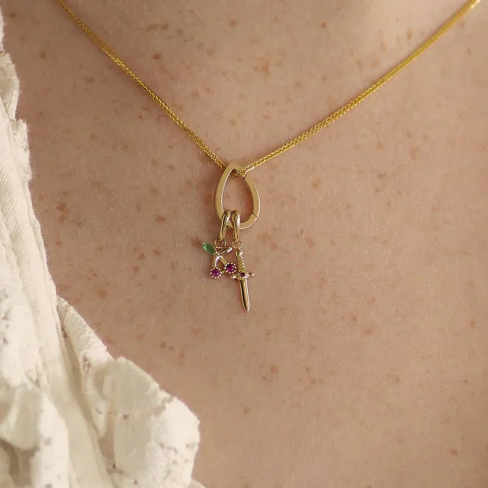 A close-up of a person's neck and chest wearing a delicate gold chain necklace with a small pendant featuring a dagger and a tiny green and purple flower charm.
