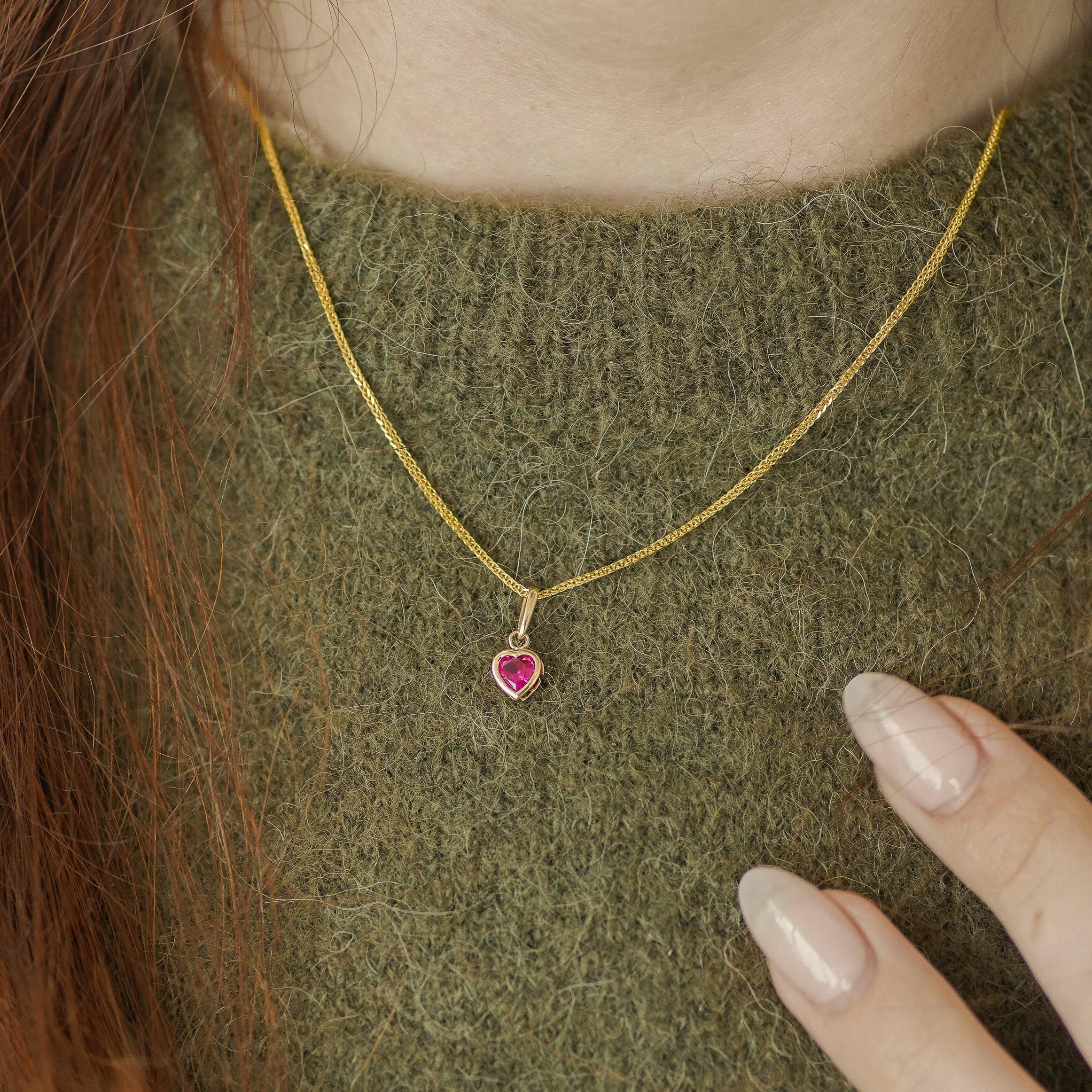 A close-up of a person wearing a delicate gold chain necklace with a small heart-shaped red gemstone pendant, styled over an olive green knit sweater.
