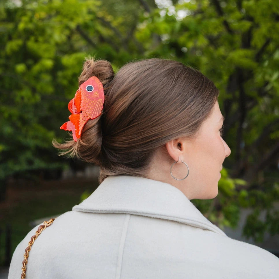 a person with brown hair and beige coat wearing a red hair clip over a green verdant background