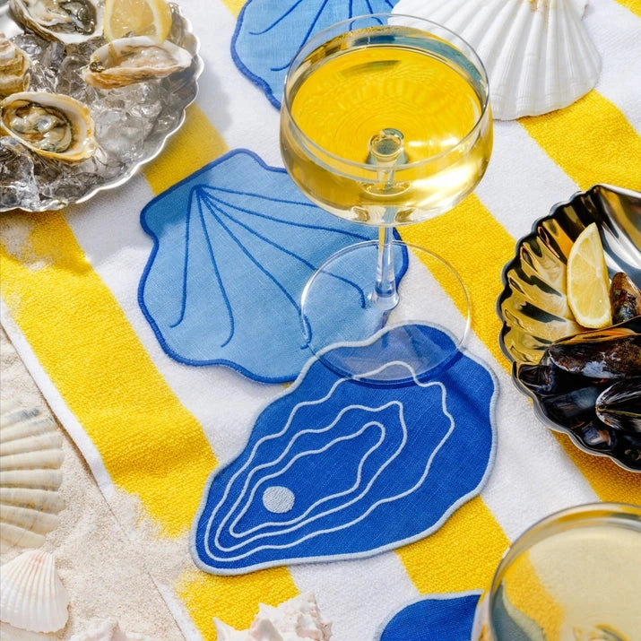 Table setting with blue shell coasters, white wine glasses, and oysters on a yellow and white checkered tablecloth.