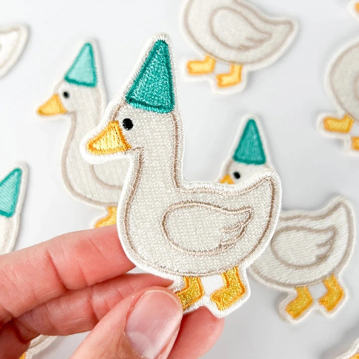 A hand holds an embroidered duck patch wearing a teal party hat and yellow beak and feet, with more matching duck patches blurred in the background on a white surface.