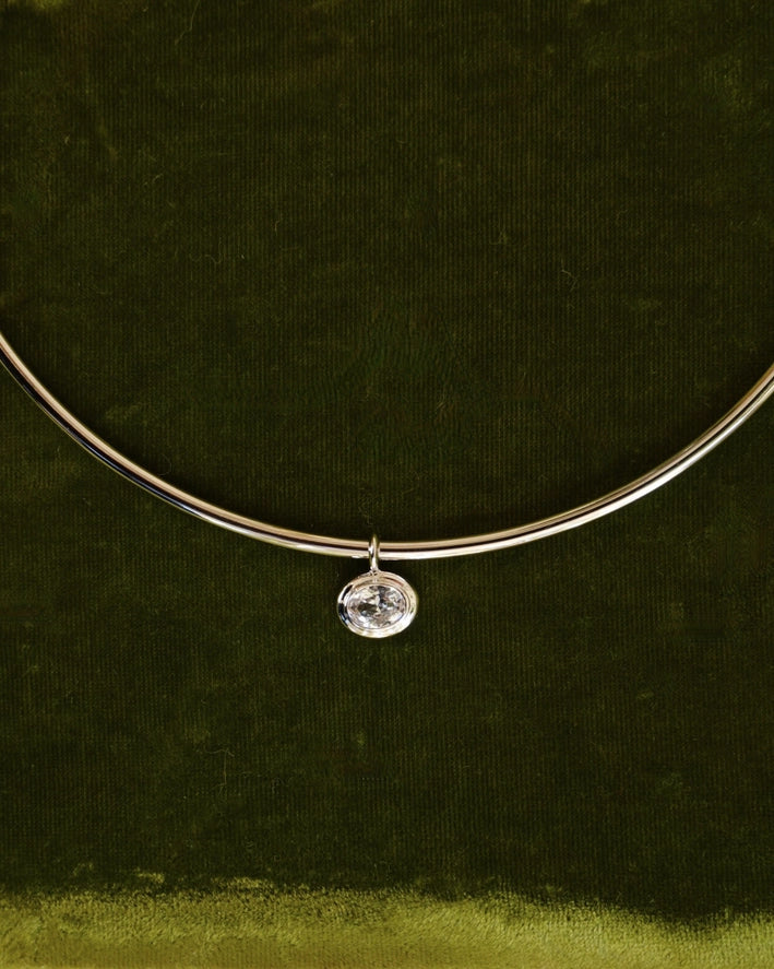a silver charm with an oval shaped bezel set white sapphire on a silver chain over a green velvet background