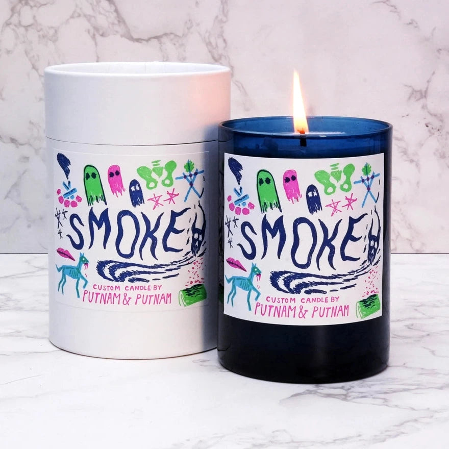 a blue candle with a white label with text reading smoke surrounded by spooky multicolored illustrations and a marble background