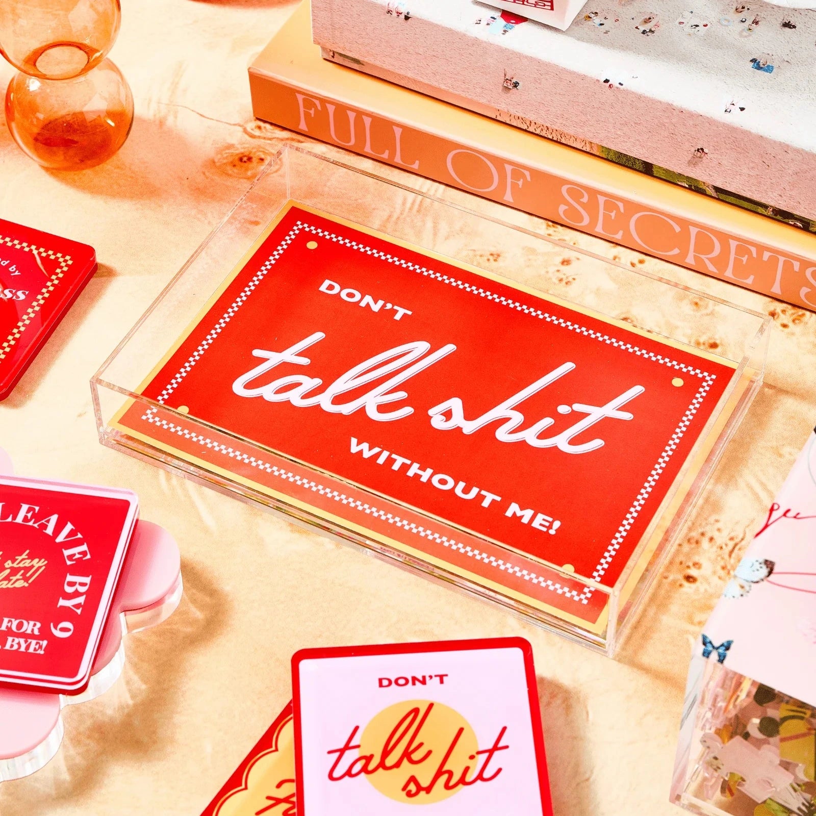 A styled flat-lay featuring a clear acrylic tray holding red cards with the phrase 'DON'T talk shit WITHOUT ME!' in white script and text. Other matching red and pink cards are partially visible around it on a warm beige surface with decorative books, glass vials, and pastel objects in the background.