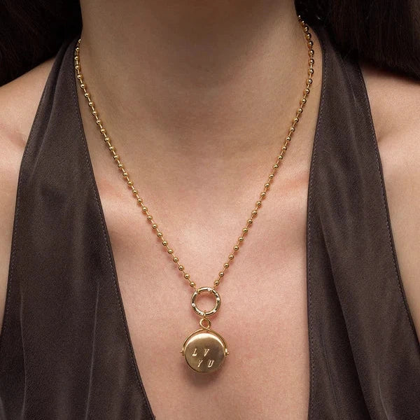 A gold necklace with a small round pendant engraved with a heart, worn by a model in a dark brown halter top.