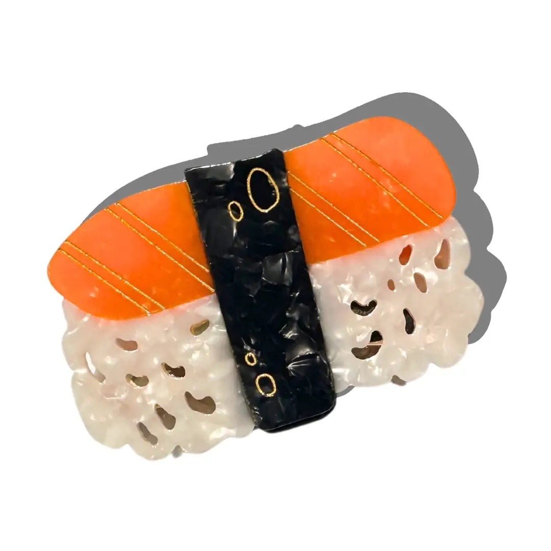 A novelty hair clip shaped like a piece of sushi, featuring orange top, a black center band with gold circular details, and a white rice base with cutout patterns, set against a plain white background with a drop shadow.