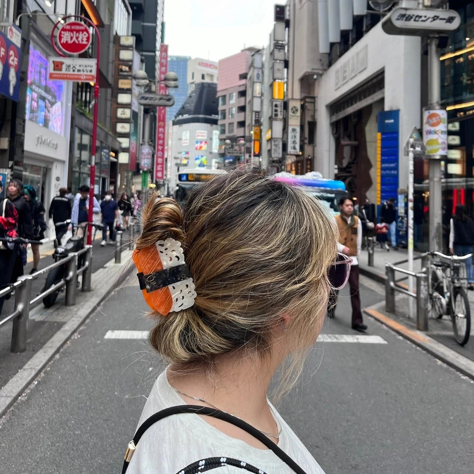 A woman in a white shirt is shown from behind in a busy city street, wearing a black and white polka dot hair claw clip with orange and white accents. The background features urban buildings, pedestrians, and street signs.