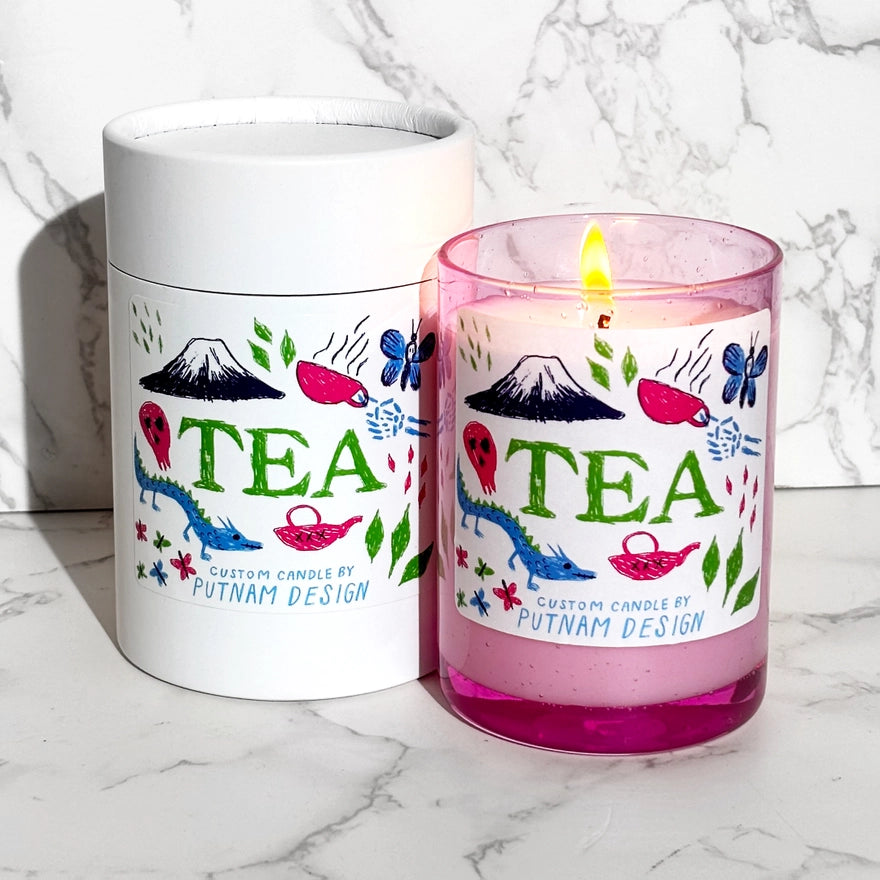 a pink candle with a white label with green text reading tea with japanese inspired drawings around over a marble background
