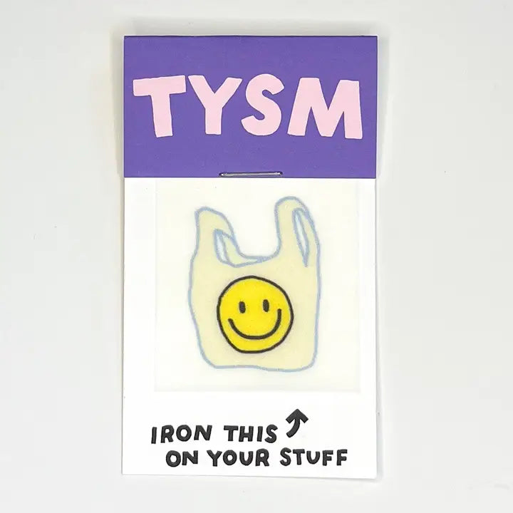 A novelty enamel pin shaped like a yellow smiley face inside a white plastic bag, mounted on its original backing card. The card features a purple top section with the word 'TYSM' in pink and a white bottom section with the text 'IRON THIS ON YOUR STUFF' and an arrow.