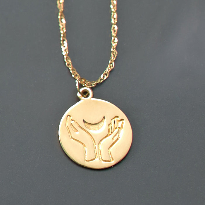 Gold necklace with a circular pendant featuring hands holding a moon design on a gray background.