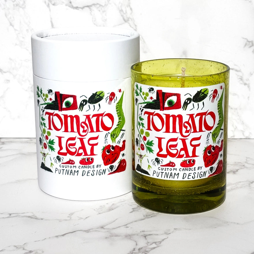a green candle with a white label with text reading tomato leaf with bug and garden motif illustrations around over a marble background