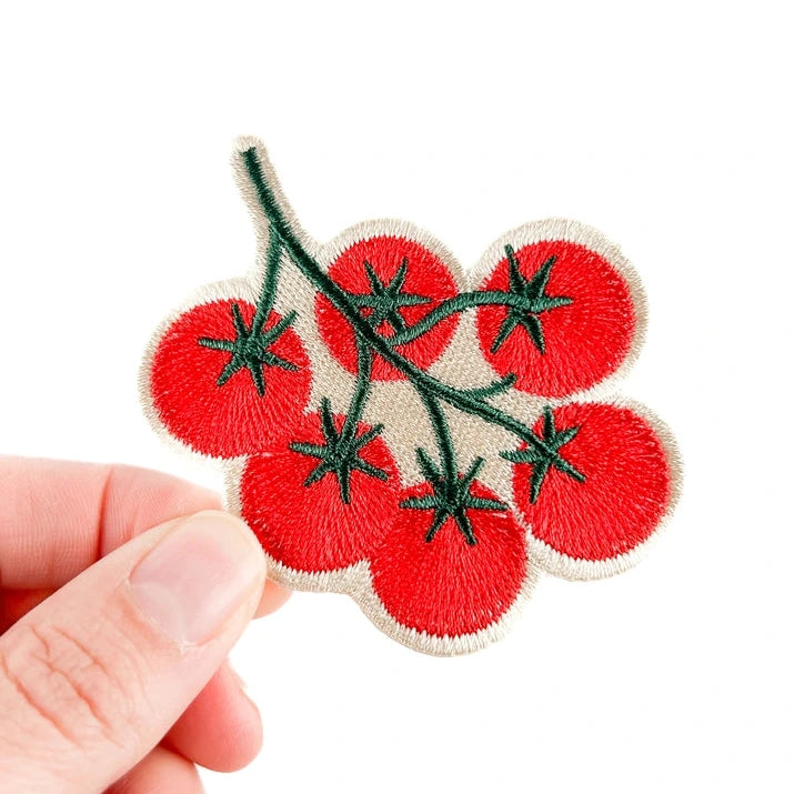 a red and green iron on patch of tomatoes on the vine held by a hand over a white background