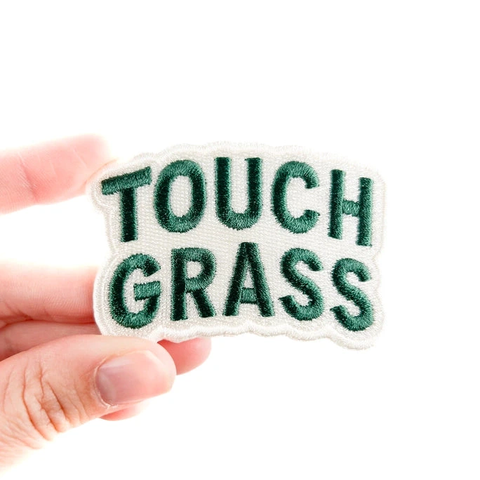 a white and green iron on patch that reads touch grass held by a hand over a white background