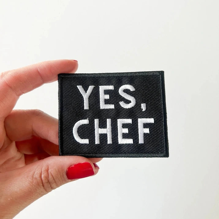 a black and white iron on patch that reads yes chef held by a hand over a white background