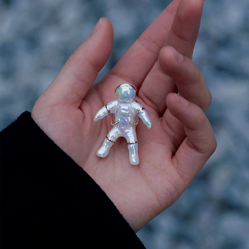a luster finished ceramic brooch in the shape of an astronaut held in a hand over a blue and back background