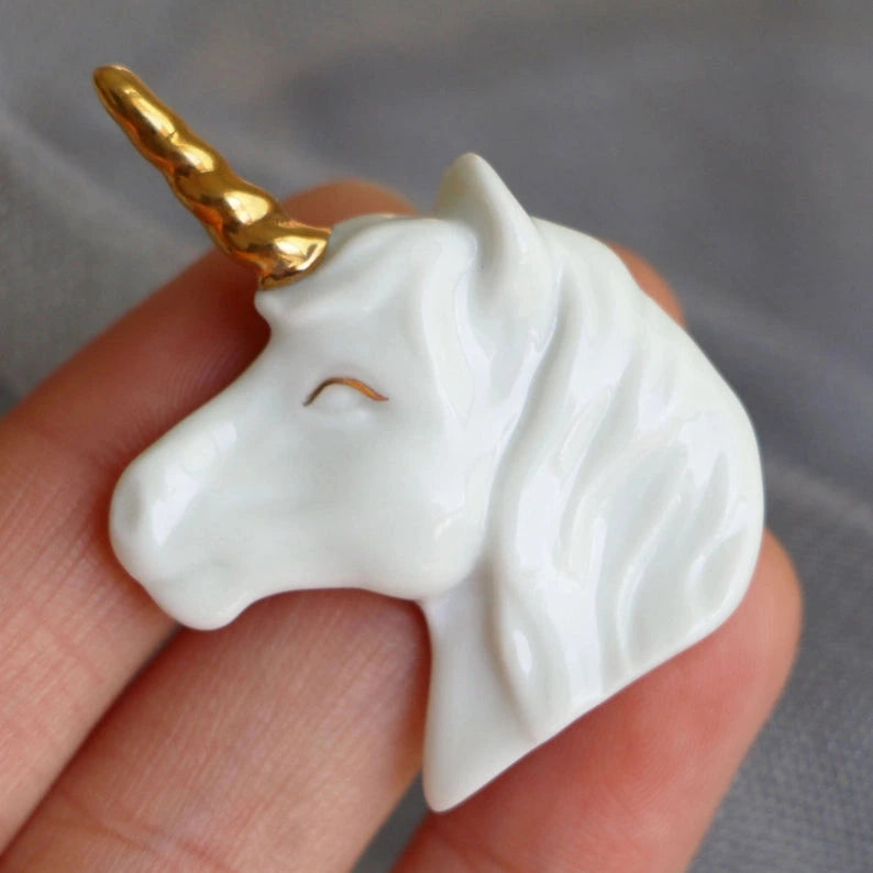 a white and gold ceramic brooch in the shape of a unicorn held by a hand over a grey background