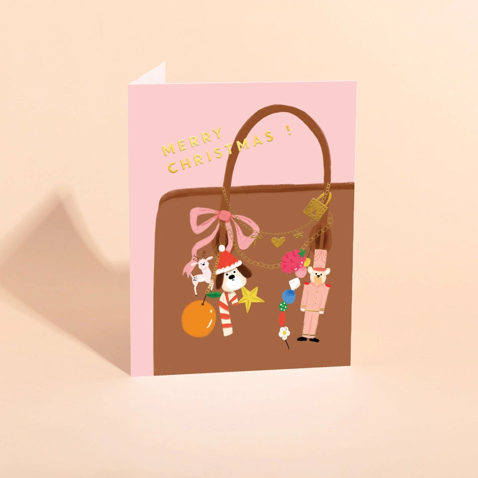 a greeting card with a pink background and an illustration of a brown handbag with a collection of colorful bag charms and text above reading merry christmas