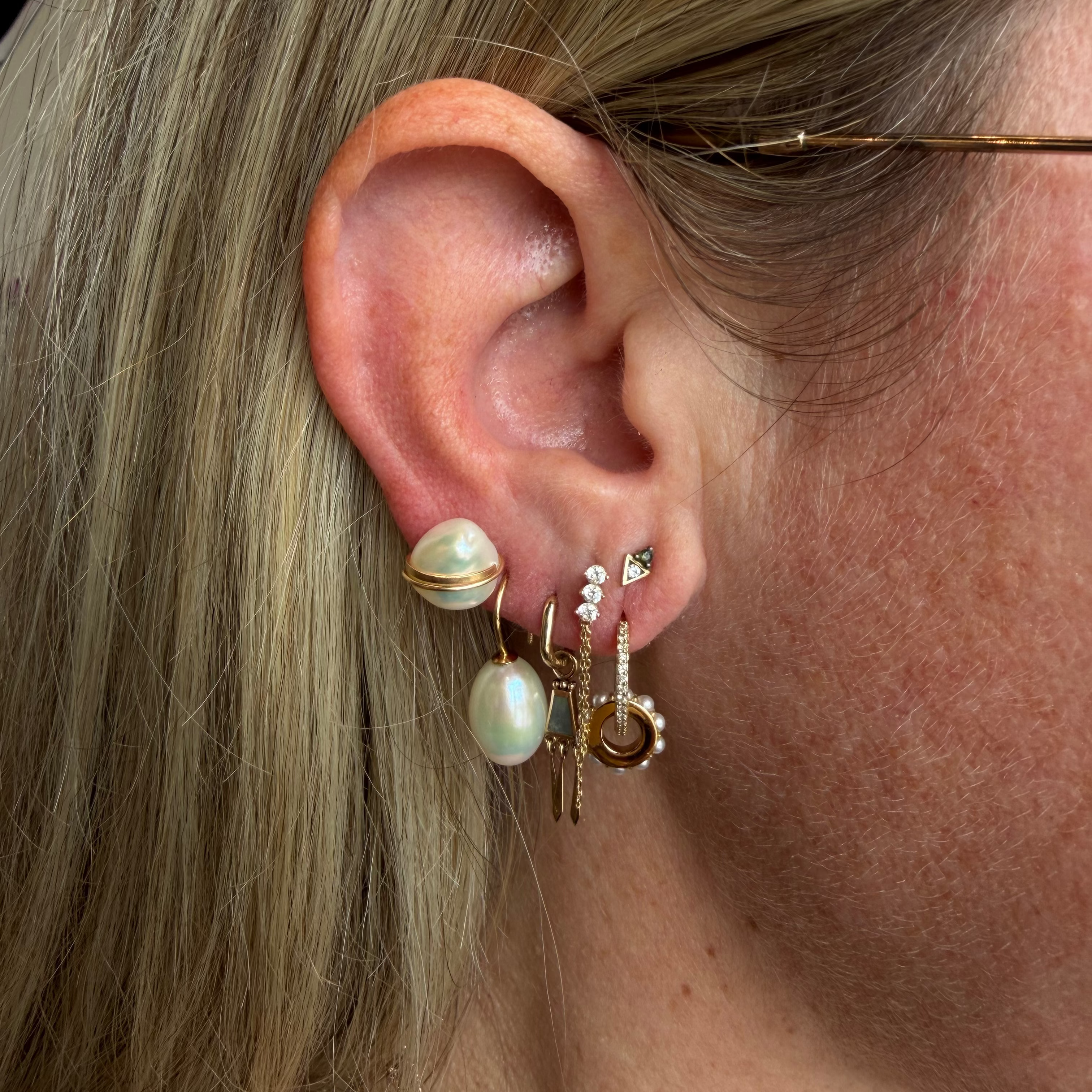 A close-up of a person's ear wearing multiple gold-toned earrings, including pearl and bead drop earrings, stud earrings, and a small hoop earring.