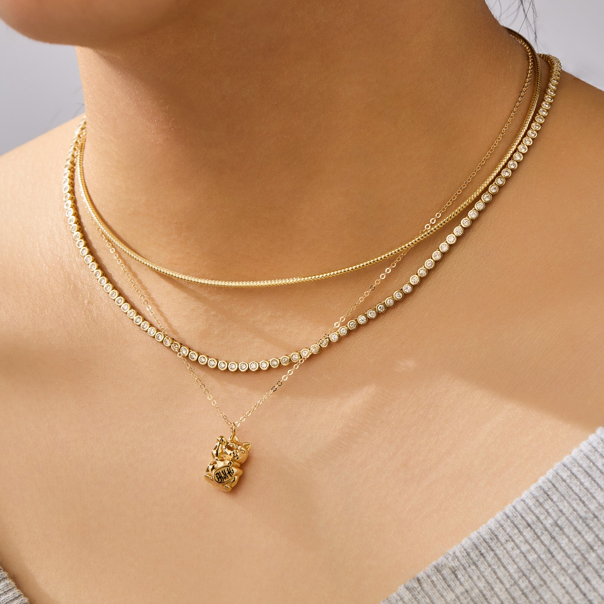 Close-up of a person wearing two gold necklaces with a lucky cat pendant on a neutral background